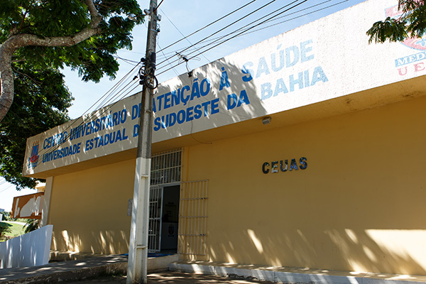 Imagem da fachada do Ceuas. Um muro de cor amarela, com um poste na frente. Acima, é possível ver um letreiro escrito, em azul, Centro Universitário de Atenção à Saúde, Universidade Estadual do Sudoste da Bahia. Na frente do módulo, existe uma porta, com portão de grade aberto.