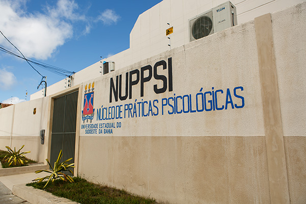 Fachada do Núcleo de Práticas Psicológicas da Uesb. Muro bege com a sigla Nuppsi e o nome do setor escritos em preto e azul.