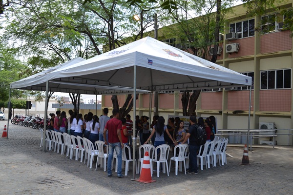 A imagem apresenta um grupo de estudantes de mãos dadas reunidas debaixo de um toldo que foi montado na parte externa da Universidade. Dois rapazes estão fora do grande círculo olhando a ação. 