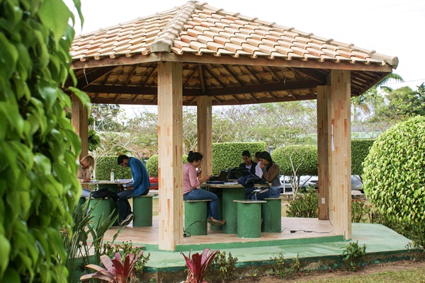 Imagem de uma área verde da Uesb, com um quiosque ao fundo e duas mesas com bancos de cimento pintados de verde. Nos bancos, alunos estão sentados e lendo.