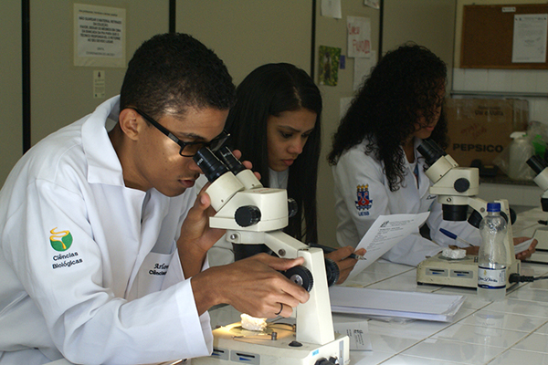 Três alunos vestidos com jalecos brancos, observam atentos em microscópios. Na manga dos jalecos é possível ler o nome do curso de "Ciências biológicas", além do brasão da Uesb.