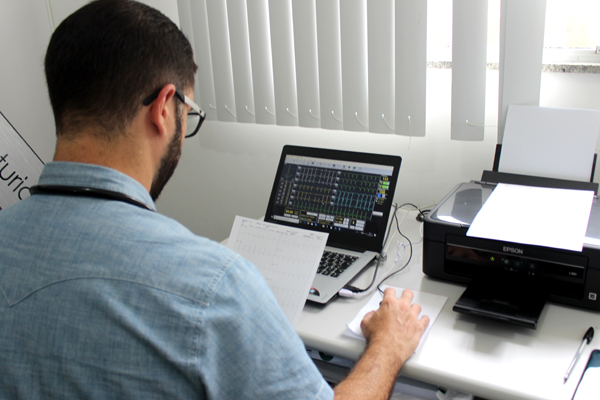 Na imagem, é possível ver um homem de costas, com camisa azul e óculos, olhando e segurando um papel e com a mão no mouse, que está conectado a um notebook. Na tela do notebook, é possível ver gráficos referentes ao exame. Na mesa onde está esse notebook, ainda é possível ver uma impressora preta.