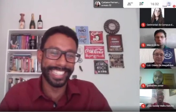 Imagem de uma tela do Google Meet, com um estudante negro, de óculos, camisa vermelha, sorrindo ao lado esquerdo e várias telas menores ao lado direito com outras cinco pessoas durante a cerimônia de colação de grau