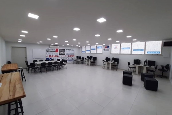 Imagem de um dos Espaços Colaborar, ambiente amplo, com muito branco e janelas ao fundo. O espaço conta com mesas, cadeiras e bancos estilo puffs