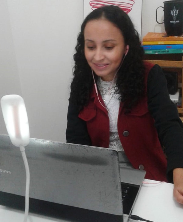 Imagem da estudante Natália Silva, em frente a um notebook prata com uma luminária branca logo atrás do notebook. Ela veste uma blusa branca, uma jaqueta vinho com mangas pretas e usa um fone de ouvido.