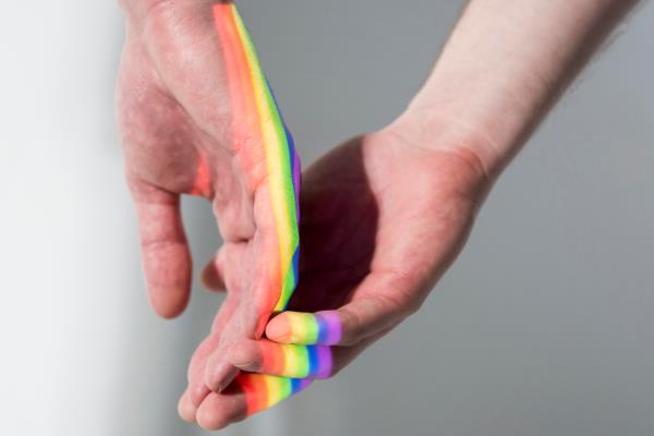 Em fundo cinza, duas mãos masculinas se sobrepõem, com o reflexo do arco-íris sobre elas.