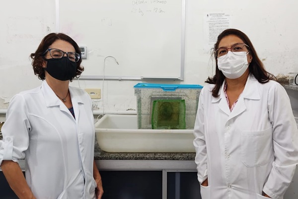 Imagem com as duas mulheres pesquisadoras responsáveis pelo estudo, ambas usando jaleco branco, óculos e máscaras. À esquerda, a professora Simone Gualberto. À direita, a professora Débora da Silva. Ao fundo, uma bancada é vista com objetos utilizados na pesquisa.