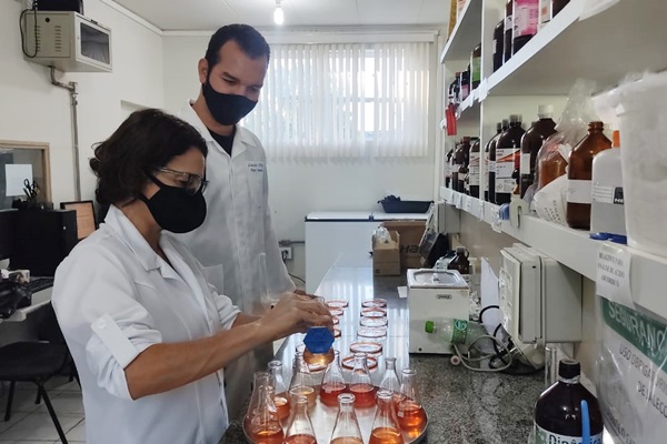 Imagem de uma mulher manuseando um vidro com solução laranja em um laboratório. Ao fundo dela, um homem, também pesquisador, acompanha o procedimento. Eles estão em uma bancada de laboratório, com vários objetos de pesquisa e vidros em cor marrom escura.