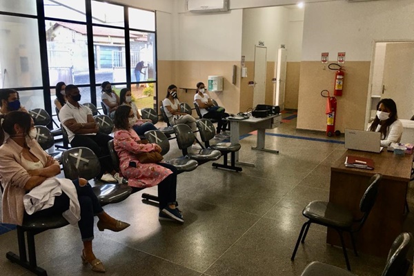 Foto da recepção da Clínica Escola de Fisioterapia, com alunos e alunas sentados distanciados em cadeiras e uma professora do outro lado do ambiente também sentada com um notebook em sua frente