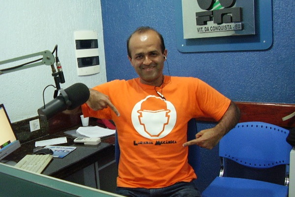 Homem sentado e sorrindo em um estúdio de rádio aponta para uma ilustração em sua camisa de cor laranja. À sua volta, estão duas bancadas, microfone, teclado, computador e papeis sobre a bancada da esquerda e, ao lado direito, uma cadeira azul de plástico vazia.