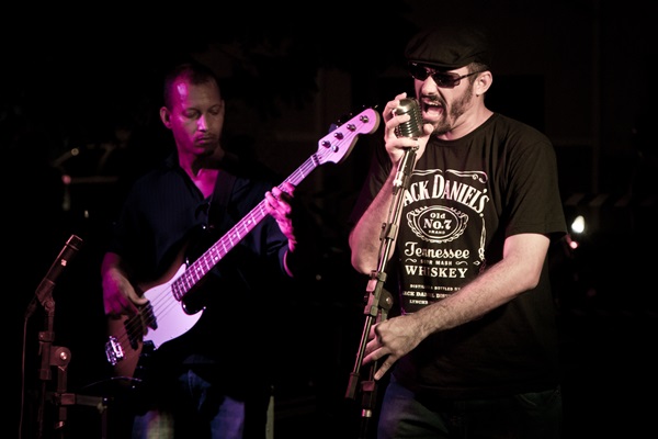 Dois homens se apresentando num palco. À esquerda, homem negro com blusa de manga comprida na cor preta e calça jeans toca baixo. Na direita, homem branco de camisa preta com dizeres na cor branca segurando o microfone com as duas mãos. Ele usa óculos escuros e boina.