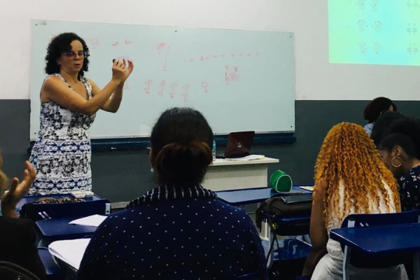 Imagem de uma sala de aula onde a professora gesticula com as mãos em Libras para os alunos que estão sentados de costas para a foto