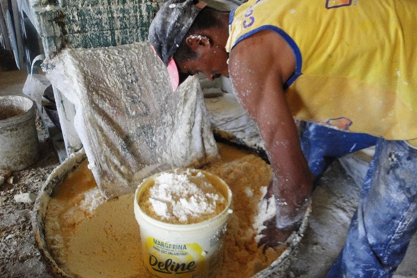 Imagem de um homem negro, com camiseta regata amarela e detalhes azuis, e uma calça azul melanda de farinha. O homem mexe em um recipiente redondo grande cheio de farinha e com um balde menor branco, no qual ele enche de farinha.