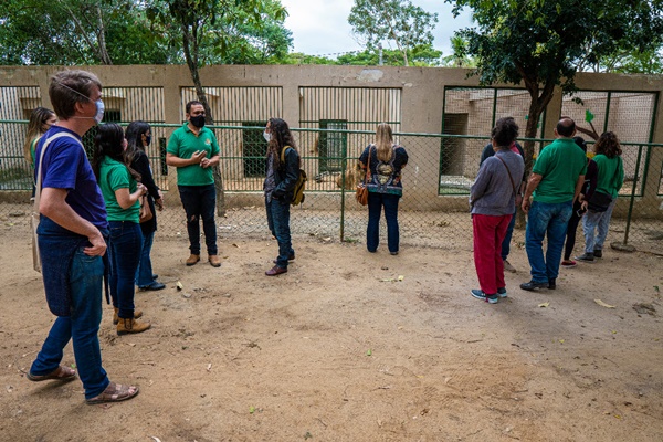 Imagem com várias pessoas em pé, conversando entre si, em frente a uma estrutura de jaulas vazias. Entre as jaulas e as pessoas, há uma proteção de metal. O ambiente está cercado de árvores e com chão de terra.