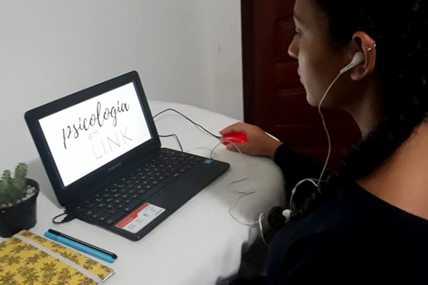 Mulher jovem de costas com notebook à frente, aberto com a marca "Psicologia em link" na tela. Ela segura o mouse e usa fones de ouvido.