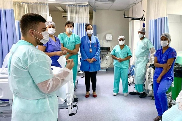 Imagem de um quarto de hospital, com profissionais de saúde todos com roupas de hospital, máscara, conversando em uma roda. Ao fundo, é possível visualizar camas de hospital