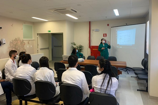 Imagem de uma sala de reunião. Ao fundo, uma mulher com máscara e jaleco verde escuro, está apresentando um trabalho exibido no projetor. Em frente a ela, diversos estudantes da Residência, com jalecos branco, de costas, assistem à apresentação.