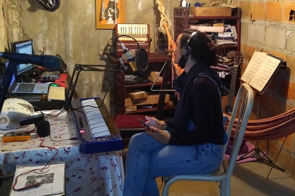 Imagem de uma mulher sentada em uma cadeira, de calça jeans e camisa preta de mangas longas, com um fone no ouvido, segurando um celular. Ao redor, vários móveis e instrumentos musicais, em um ambiente rústico. Em frente a mulher, é possível ver um teclado e um microfone. Ela está em um estúdio.