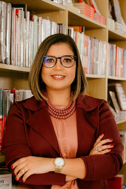 Mulher branca de cabelo castanho na altura dos ombros, que usa óculos oval grande preto com detalhes marrom, veste blusa na cor salmão com colar de bolinhas com quatros voltas num tom de cor semelhante ao da blusa e por cima um casaco na cor marsala. Ela está em frente a uma estante de livros.
