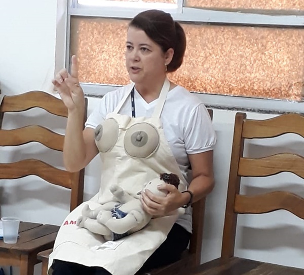 Imagem de uma mulher sentada, vestindo um avental com seios para demonstração da amamentação. No colo, a mulher segura uma boneca que simboliza um bebê. A mulher está com o dedo levantado e falando.