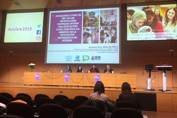 Na imagem, o professor Jonson e mais três pessoas estão compondo a mesa de apresentação do evento. Na parede, há três telões. No maior e centralizado, aparece a capa de apresentação da pesquisa do professor.