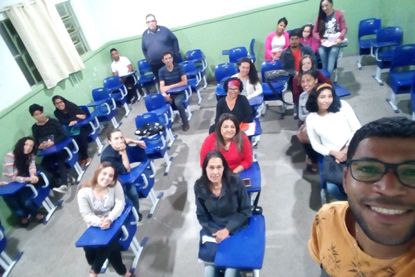 Na imagem, o professor Reinaldo aparece em pé, em primeiro plano, fazendo uma foto com a câmera frontal (estilo selfie) e no fundo, os alunos estão sentados na carteira sorrindo para a foto, dois alunos ao fundo, estão de pé. 