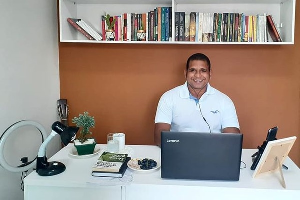 O professor Wesley Amaral está sorridente, numa mesa de escritório branca, em frente ao notebook, e atrás dele é possível ver uma estante com vários livros