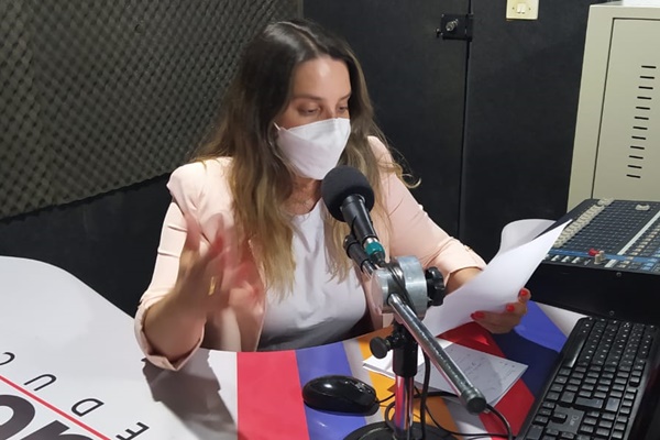 Aline Ferraz usando máscara, sentada diante do microfone enquanto lê um papel, no estúdio da Uesb Fm