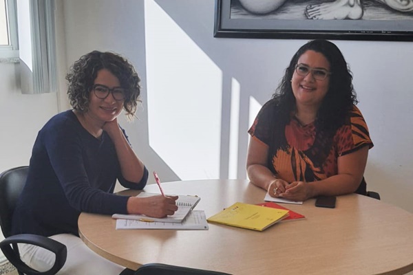 Duas mulheres sentadas à mesa rindo, sobre a mesa alguns papeis 