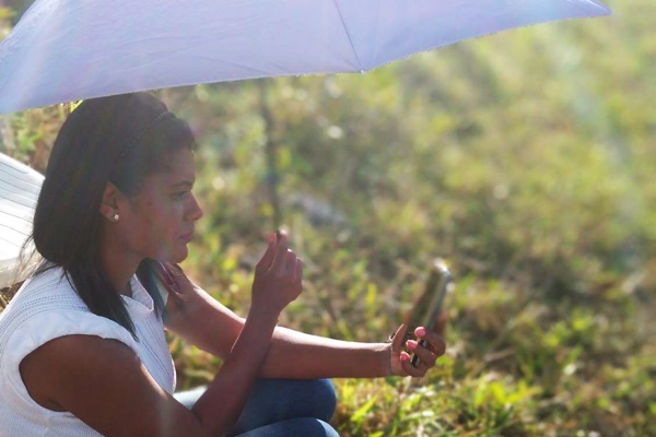 Mulher agachada em meio a vegetação, segurando um guarda chuva com uma das mãos e na outra segurando um celular.