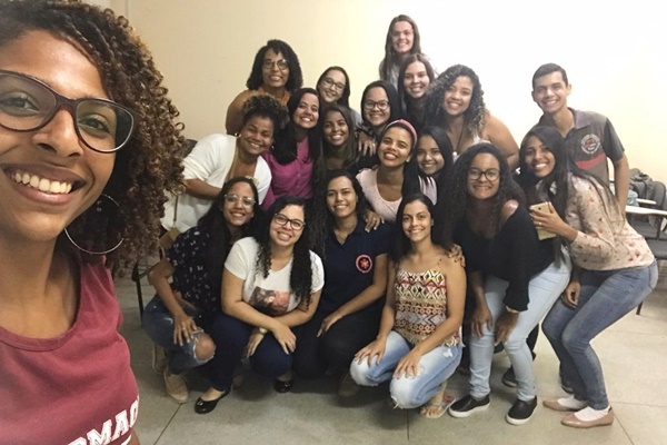 Professora negra de cabelos cacheados tirando foto no estilo de selfie com a turma de alunos do curso de Enfermagem que estão num grupo de 18 pessoas na sala de aula.