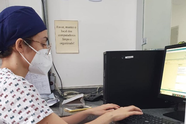 Mulher branca jovem usando touca médica azul, máscara e óculos em frente a tela de um computador digitando no teclado.
