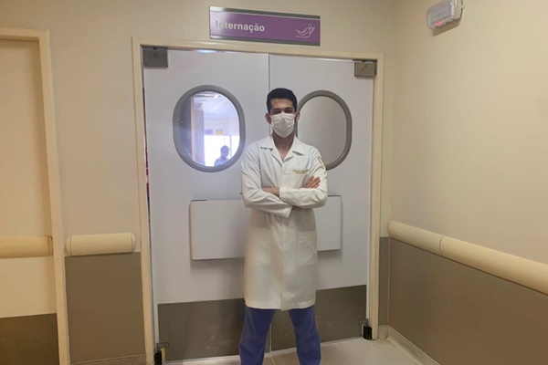 Homem branco, alto, de cabelo preto, usando jaleco branco e calça azul, na porta do setor de internação de um hospital.