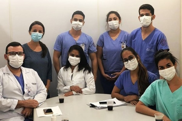 Grupo com oito pessoas, quatro em pé e quatro sentadas, em volta de uma mesa numa sala.