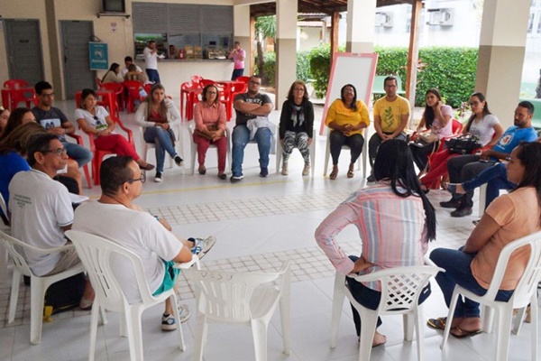 Na imagem é possível visualizar um grupo de pelo menos dezoito pessoas sentadas em círculo. Elas parecem estar direcionando o olhar e escutando uma pessoa falar. 