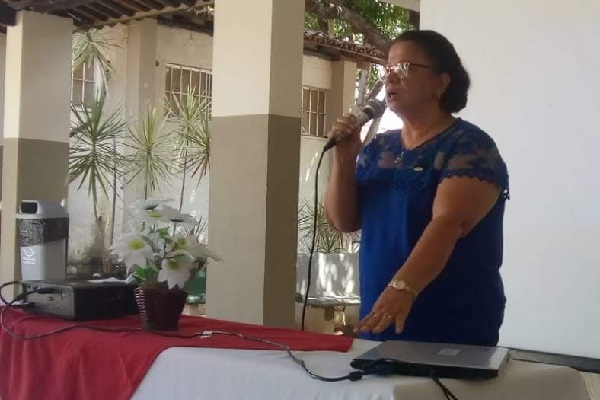 Vilma está em pé, de blusa azul. Ela segura microfone e palestra diante de uma mesa montada para a realização de evento. 