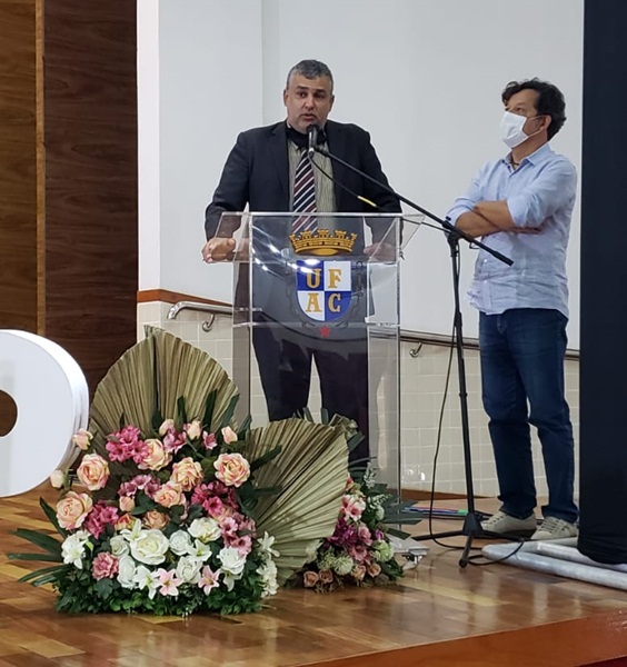 Na imagem, vemos o pró-reitor Robério Rodrigues atrás de um púlpito de transparente, com um brasão na frente. Ele está falando em um microfone de pé. Ao lado dele, está um homem, de máscara, e camisa azul clara.