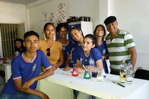 Na foto, é possível ver sete pessoas ao redor de uma bancada com vidraria de química e garrafas plásticas. Entre as pessoas, estão dois meninos estudantes, quatro meninas estudantes e o professor. Ao fundo, é possível ver armários, objetos da química e outra estudante.