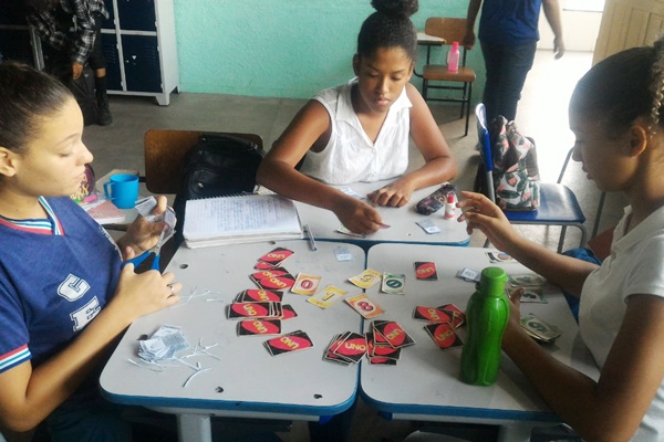 Na imagem, três estudantes estão sentadas em três carteiras de escola encostadas uma na outra. Elas estão olhando para a mesa, onde possui um jogo de cartas espalhadas, algumas viradas para baixo e outras viradas para cima.