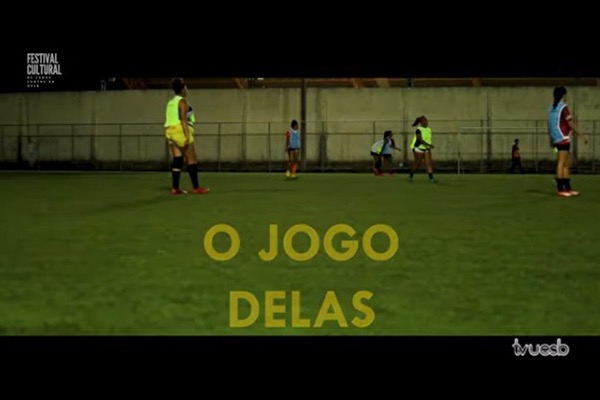Trecho da cena fílmica de "O jogo delas". Tela com a imagem de um campo de futebol com mulheres jogadoras