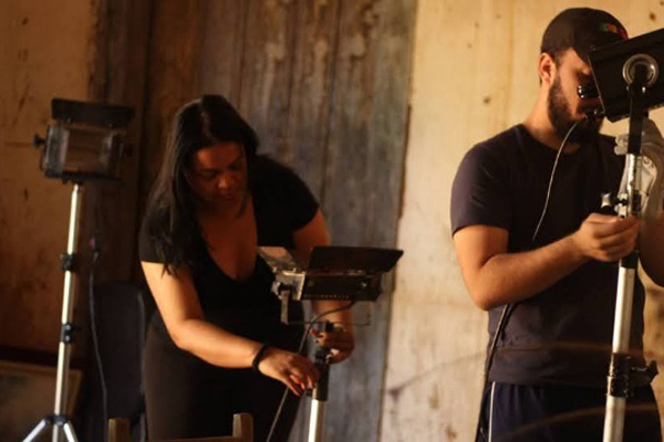 Imagem com os bastidores da produção de um TCC do curso de Cinema e Audiovisual da Uesb. Na imagem há um homem e uma mulher ajustando câmeras e aparelhos de iluminação