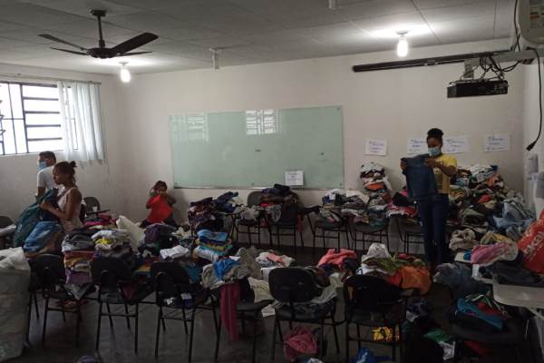 Roupas que foram doadas em cima das cadeiras numa sala de aula da Uesb. Quatros pessoas aparecem separando e dobrando as peças.