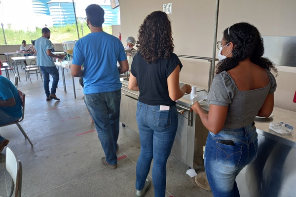 Estudantes de costas e em fila seguram o prato para colocar a refeição.